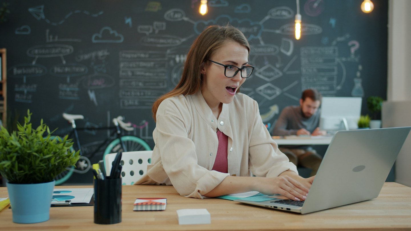 Woman with glasses excitedly works on a laptop at a desk.