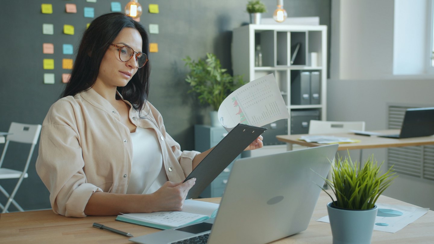 Woman with glasses reviews charts on clipboard at desk.
